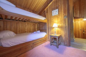 Quarto de cabana de madeira com beliche, uma mesinha com luminária e porta de madeira.