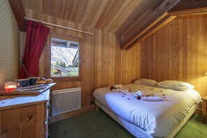 Quarto de cabana de madeira com uma cama, janela com vista para montanhas, uma mesinha com uma vela e um radiador.