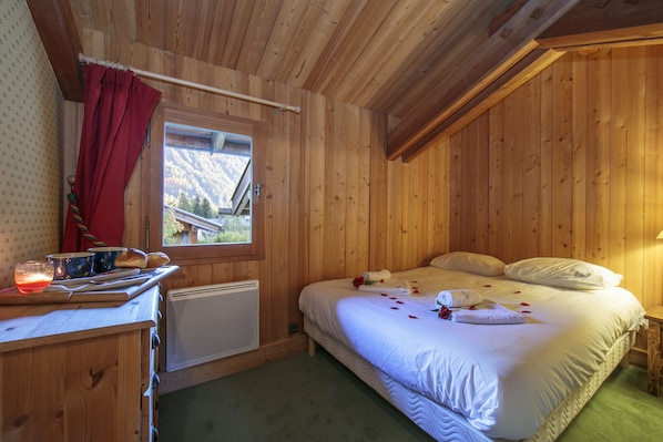 A wooden cabin bedroom with a bed, a window with a view of mountains, a small table with a candle, and a radiator.