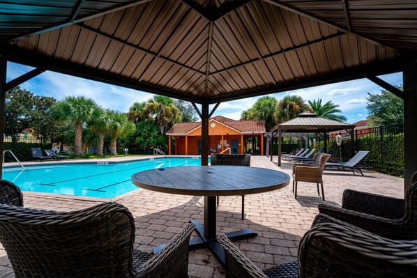 An outdoor pool area with a covered seating area, lounge chairs, and a building with a red roof in the background.