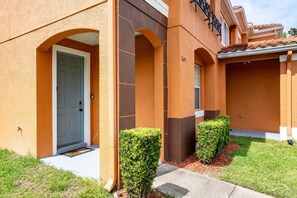A building with a beige exterior, a grey door, and a small front yard with trimmed bushes.