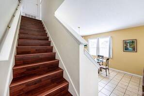 A wooden staircase leading to a room with a dining area, a painting on the wall, and a window with blinds.