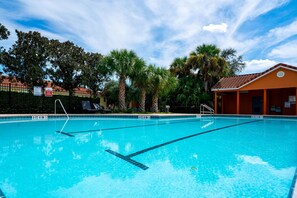 A clear swimming pool with lane dividers, surrounded by palm trees and a building with a tiled roof.