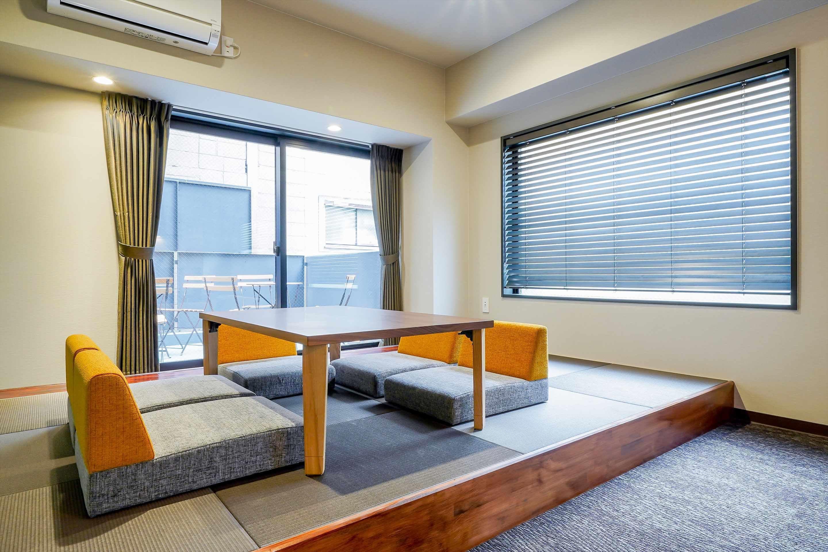 A room with a wooden table, grey and orange cushioned seating, a large window with blinds, and a view of buildings outside.