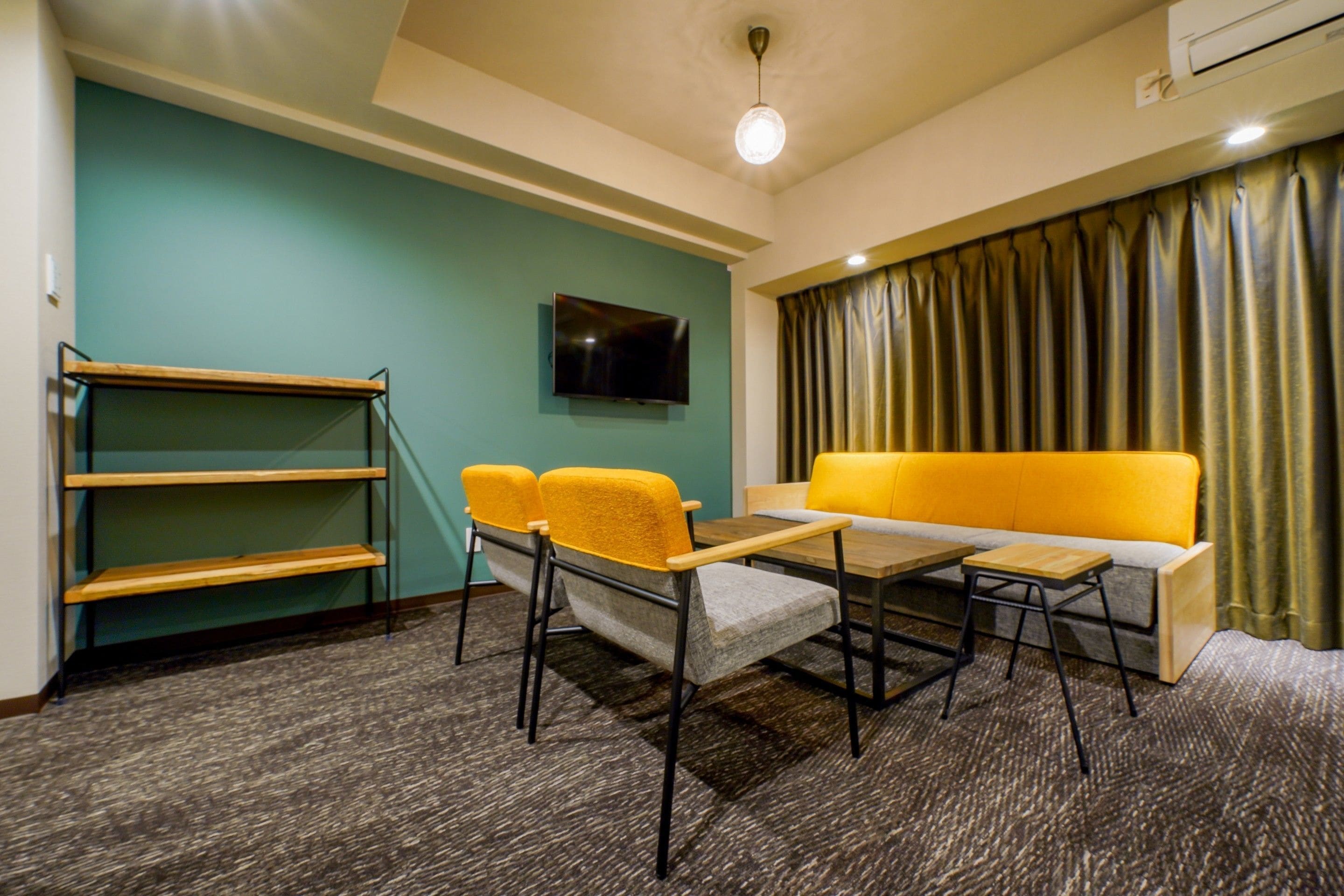 Ein Hotelzimmer mit einer türkisblauen Wand, einem Flachbildfernseher über einem Holztisch, einem gelben Sofa und einer Holzregaleinrichtung.