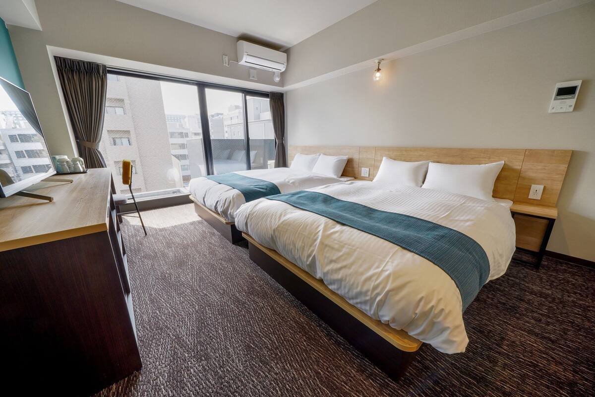 A hotel room with a large bed, a desk with a computer, a window with curtains, and a view of buildings outside.