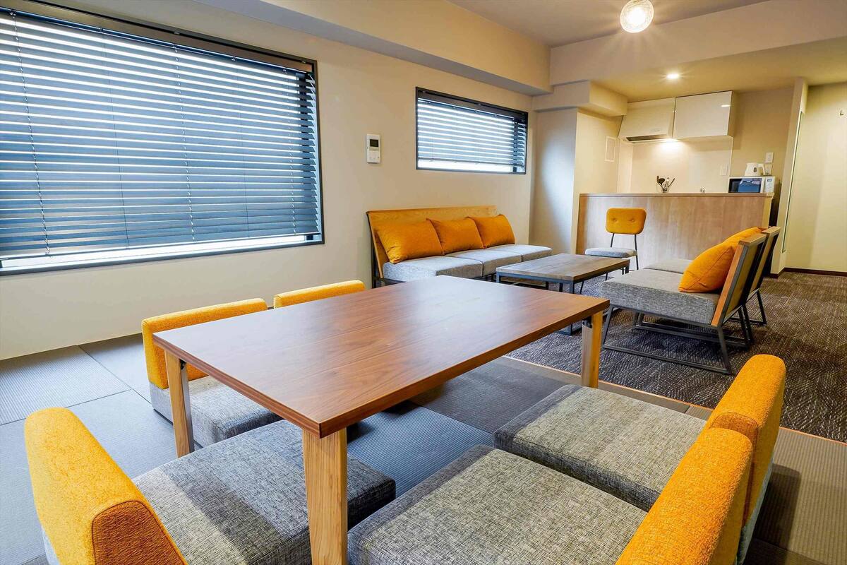 A modern hotel room with a wooden dining table, cushioned seating, and a kitchenette in the background.
