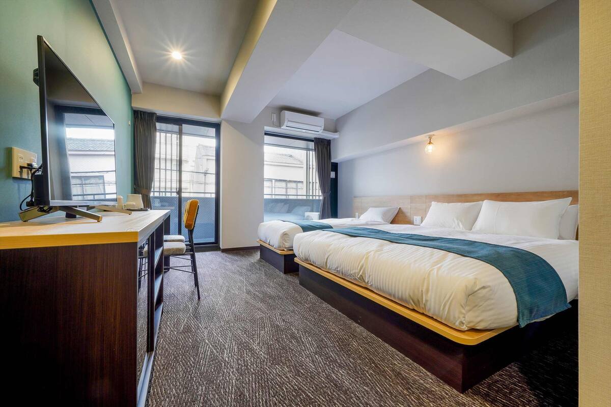 A hotel room with a desk, two beds, a chair, a large window, and an air conditioning unit.