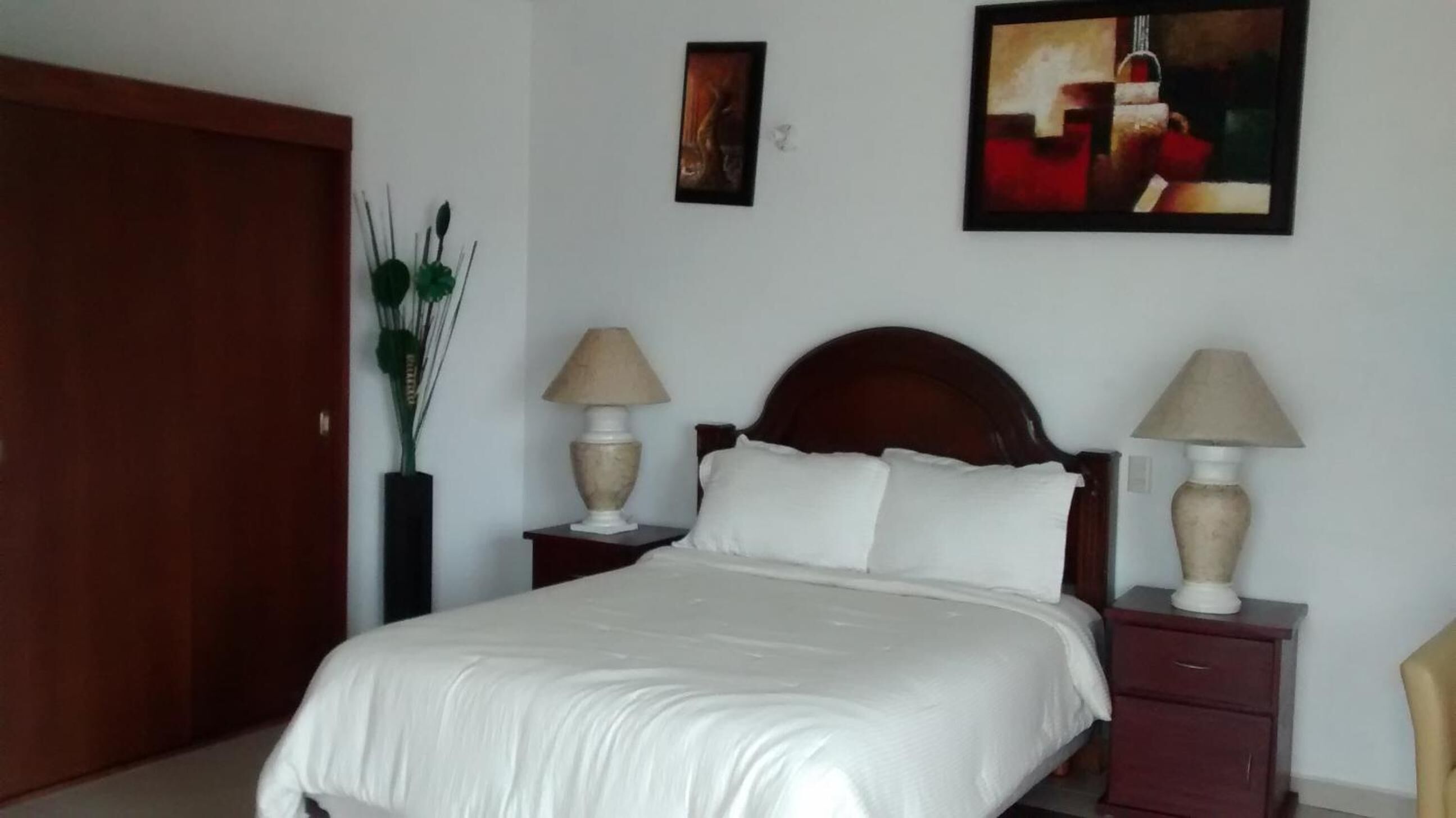 A neatly made bed with white linens, two bedside tables with lamps, a wooden door, and two wall-mounted paintings.
