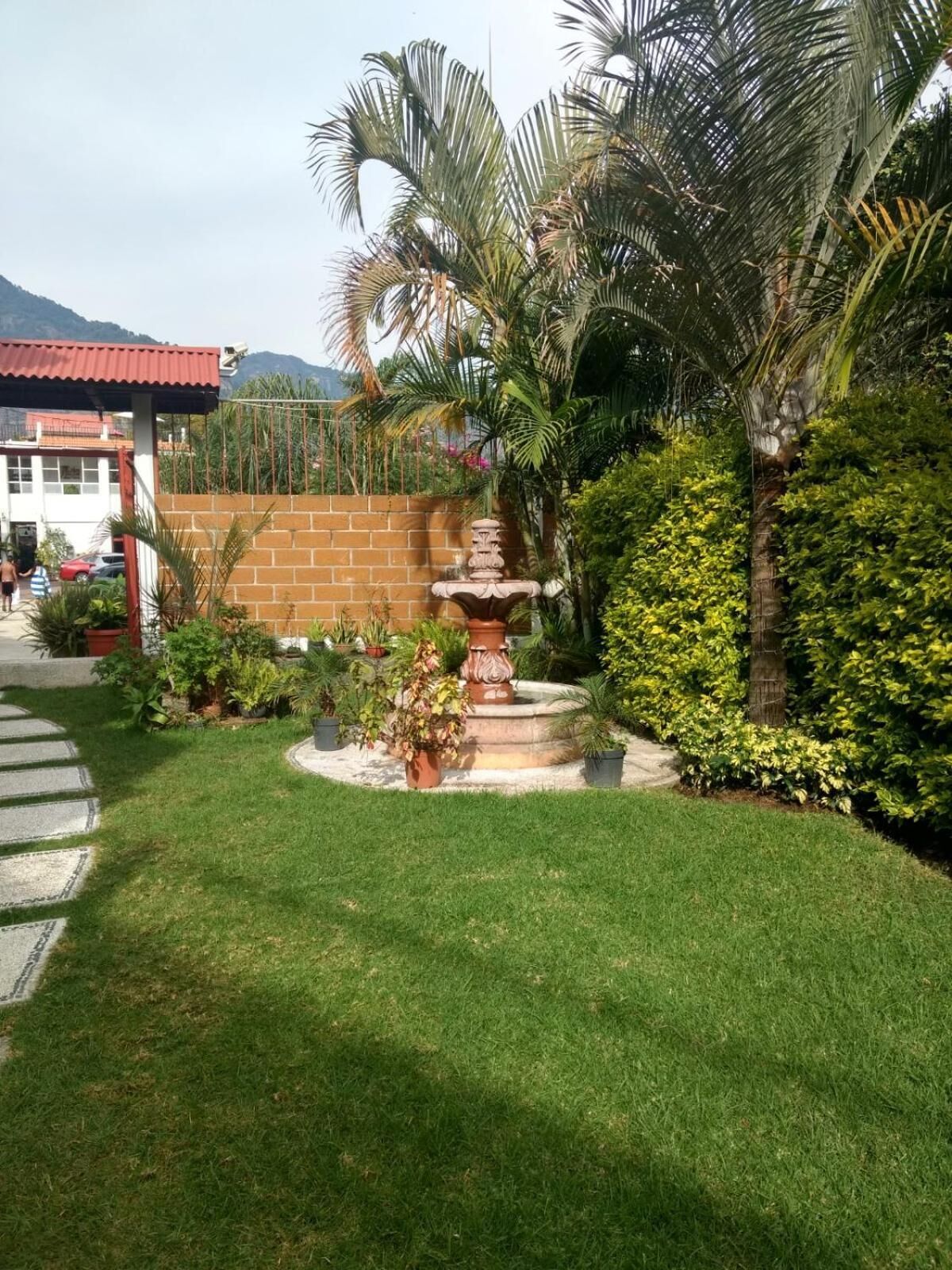 Un jardin bien entretenu, avec une fontaine centrale, entouré de différentes plantes et délimité par un mur de briques.