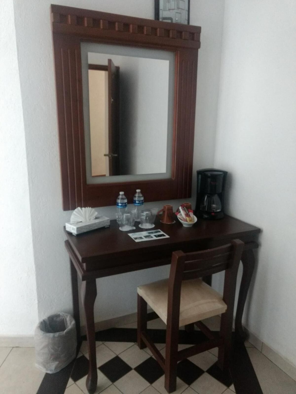 Un escritorio de madera oscura pequeño con un espejo encima, una silla, un cubo de basura y una cafetera.