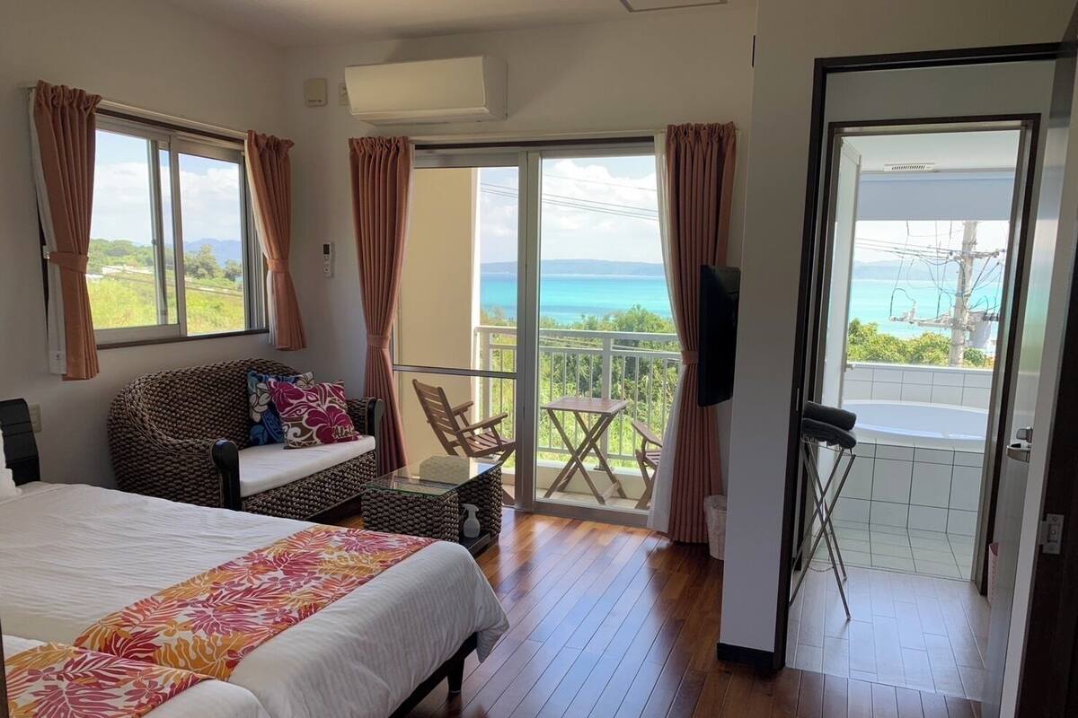 A bedroom with a bed, a sofa, a balcony with a table and chairs, and a view of the sea.