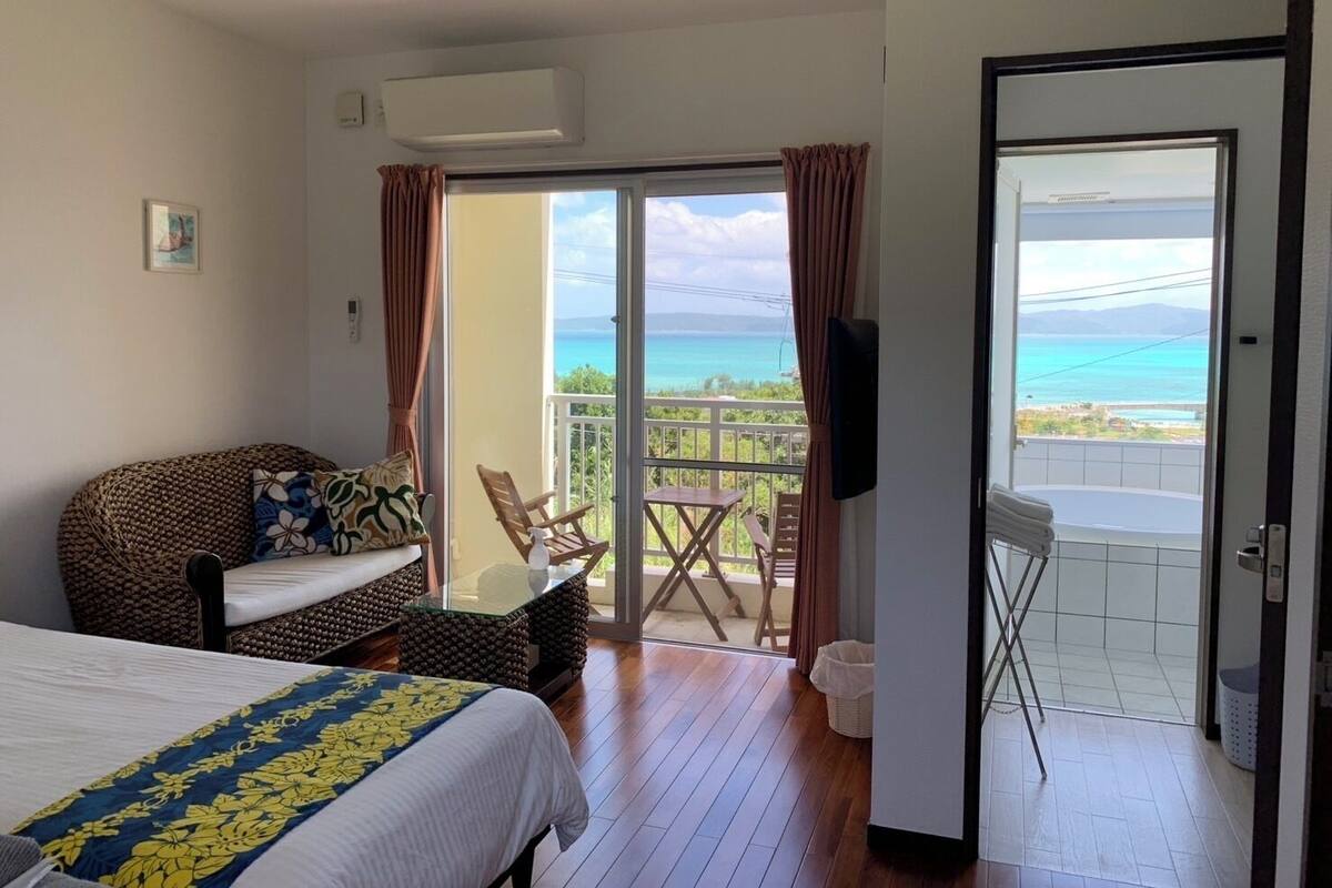 A hotel room with a bed, a sofa, a table, and a balcony with a view of the sea.