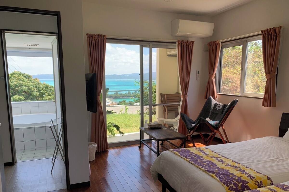 A bedroom with a bed, a chair, a TV, and a balcony with a view of the sea.
