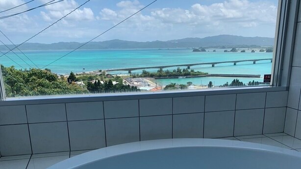 A bathroom with a bathtub, a window with a view of a turquoise sea and distant mountains. - Seabreeze Kouri (Nakijin)