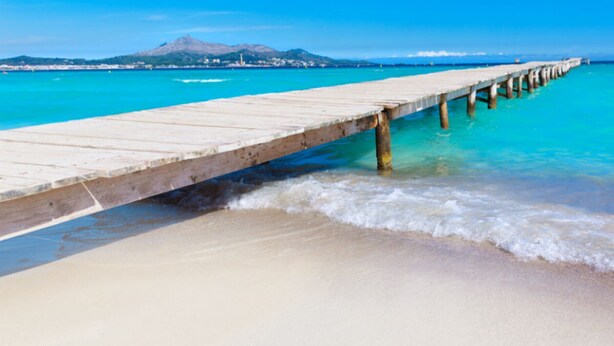 Une jetée en bois s’étendant dans des eaux turquoise, avec une plage de sable au premier plan.