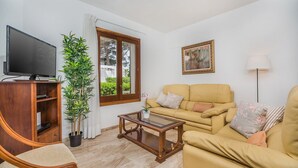 Un salon avec un canapé, une table basse, un téléviseur posé sur un meuble en bois, une plante en pot et une fenêtre avec des rideaux.