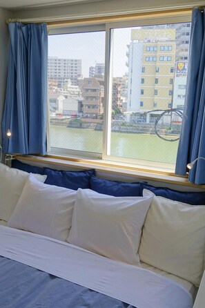 A hotel room with a bed, a window with blue curtains, and a view of buildings and a water body outside. - Fukuoka Tabiji Hostel (Fukuoka)