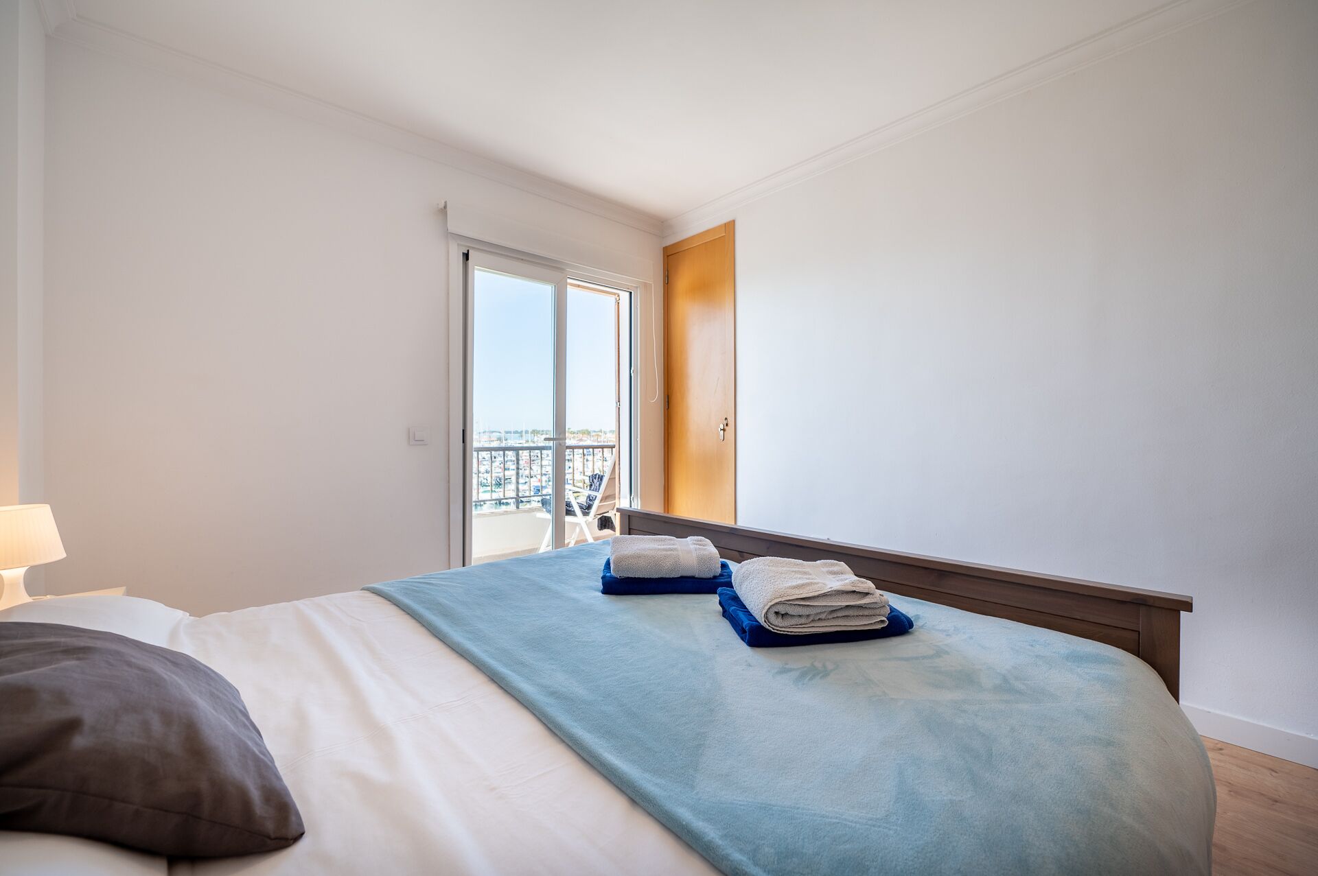 Een hotelkamer met een bed, nachtkastje, lamp en uitzicht op een jachthaven door het raam.