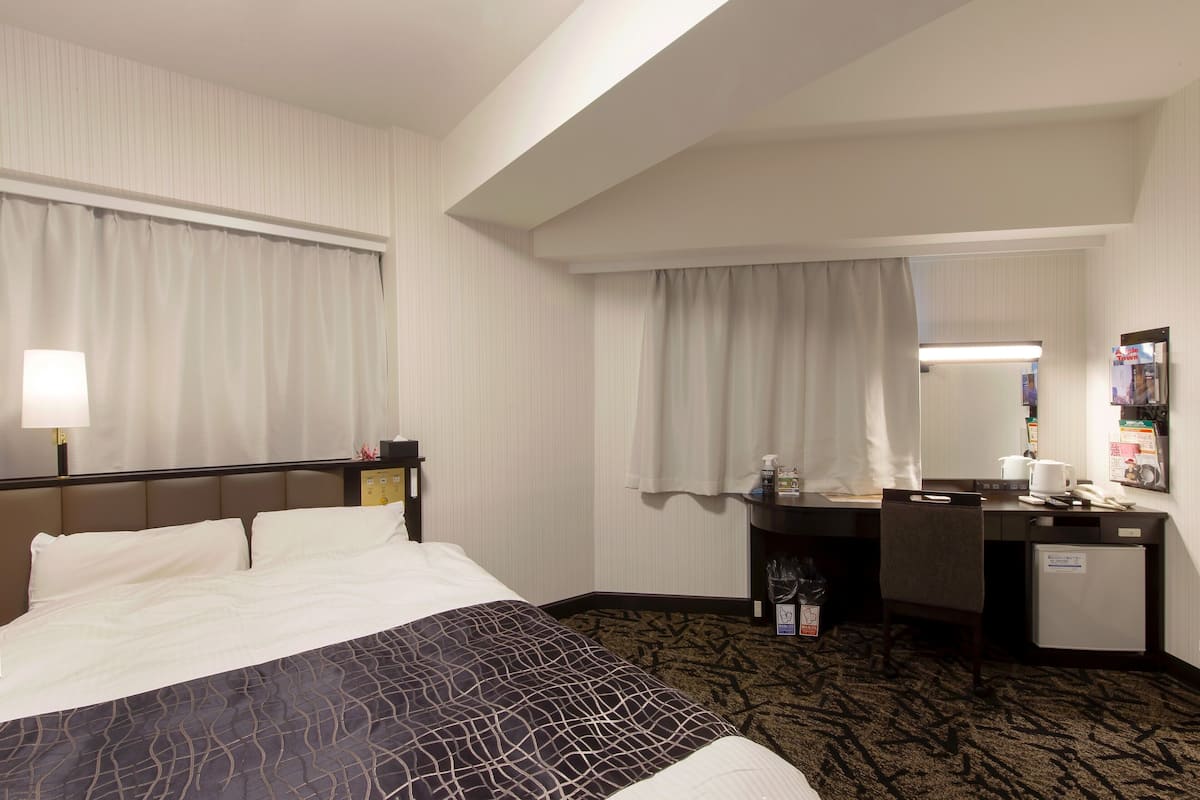 A hotel room with a bed, a desk, a chair, a small refrigerator, and a mirror.