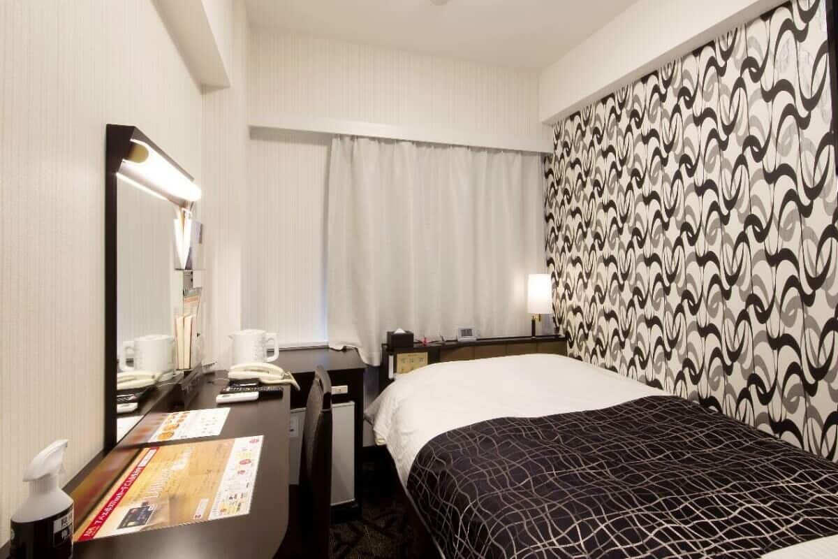 A hotel room with a bed, a desk with a telephone, a mirror, a nightstand with a lamp, and a patterned wall.