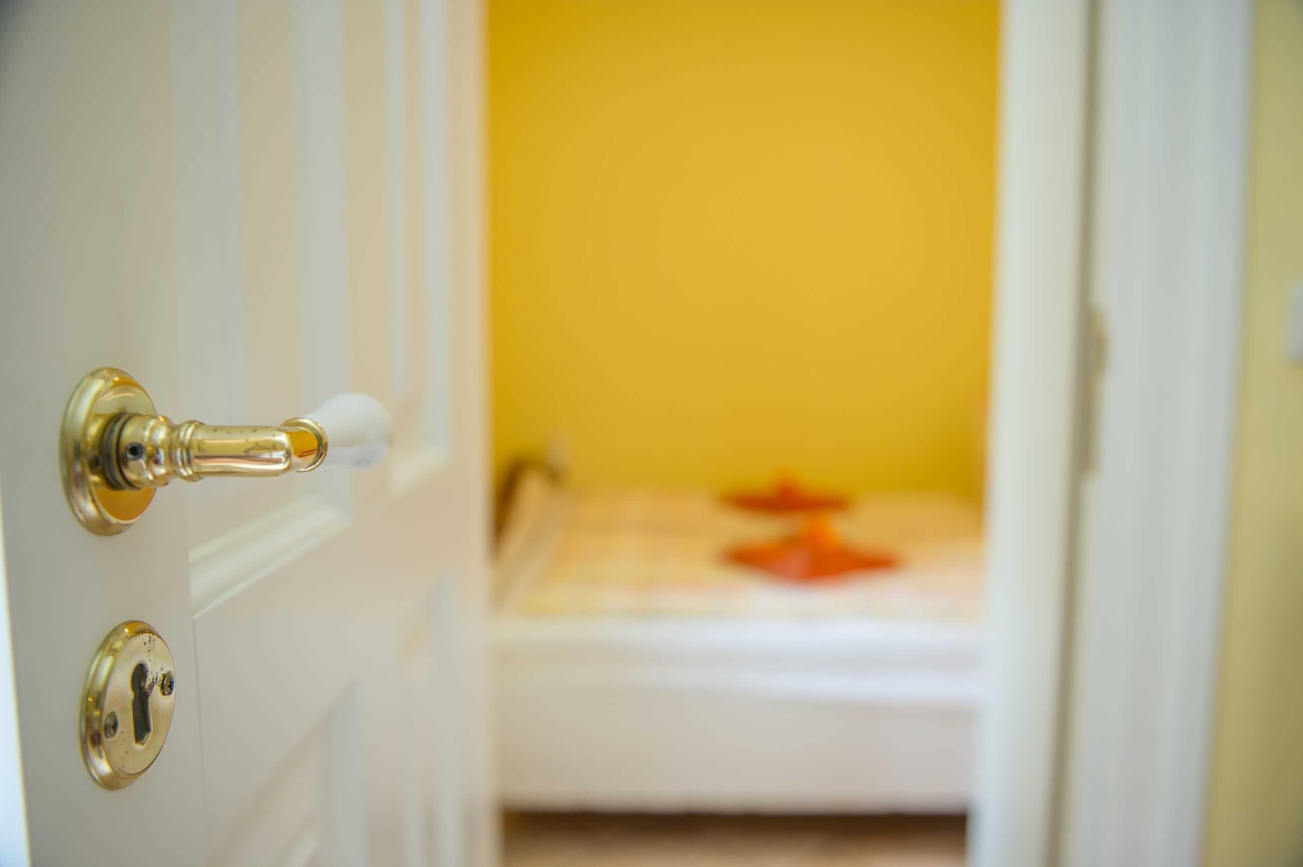 Une porte dotée d’un trou de serrure et d’une serrure, qui donne sur une pièce dont un mur est jaune et où se trouve un objet blanc sur le sol.