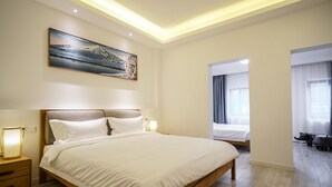 Une chambre d’hôtel moderne avec un grand lit, des lampes de chevet, un tableau au mur et une vue sur la pièce à travers une porte ouverte.
