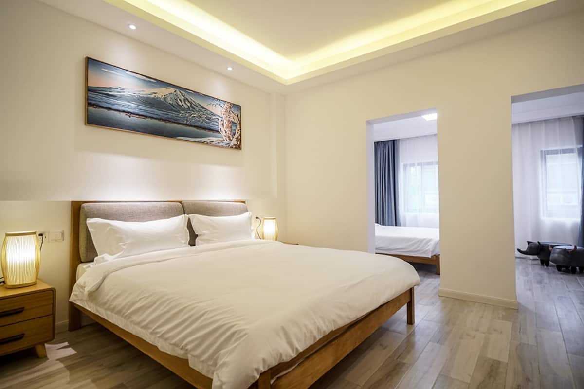 Una moderna camera d'hotel con un letto grande, lampade ai comodini, un quadro appeso e vista sull'ambiente attraverso una porta aperta.