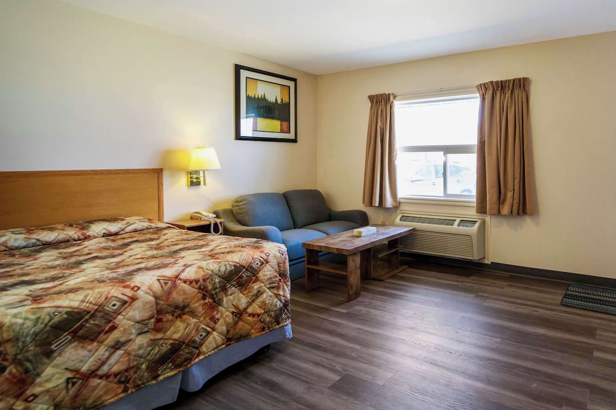 Una habitación de hotel con cama, sofá, mesa, lámpara, un cuadro en la pared, ventana con cortinas y equipo de aire acondicionado.