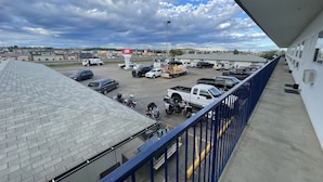 En parkeringsplats med olika typer av fordon, inklusive bilar och lastbilar, och en byggnad med ett blått räcke i förgrunden.