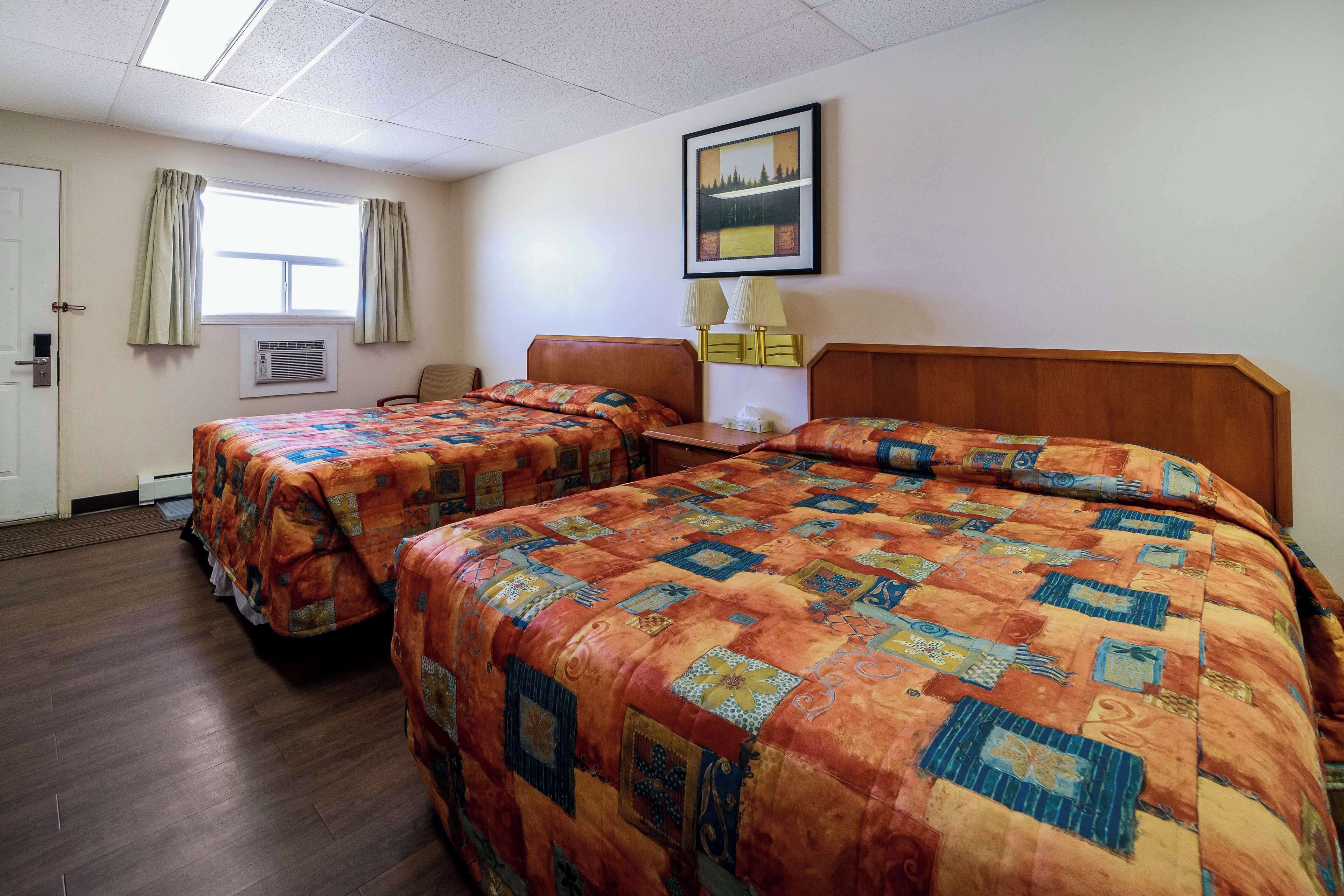 Une chambre d’hôtel avec deux lits, un bureau, une chaise, une fenêtre avec des rideaux, un appareil de climatisation et un tableau encadré au mur.