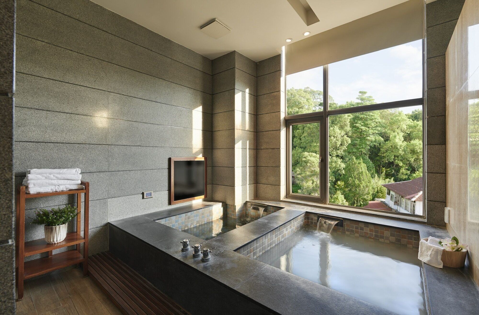 A modern bathroom with a large bathtub, a wooden shelf with towels, and a view of trees through a large window.