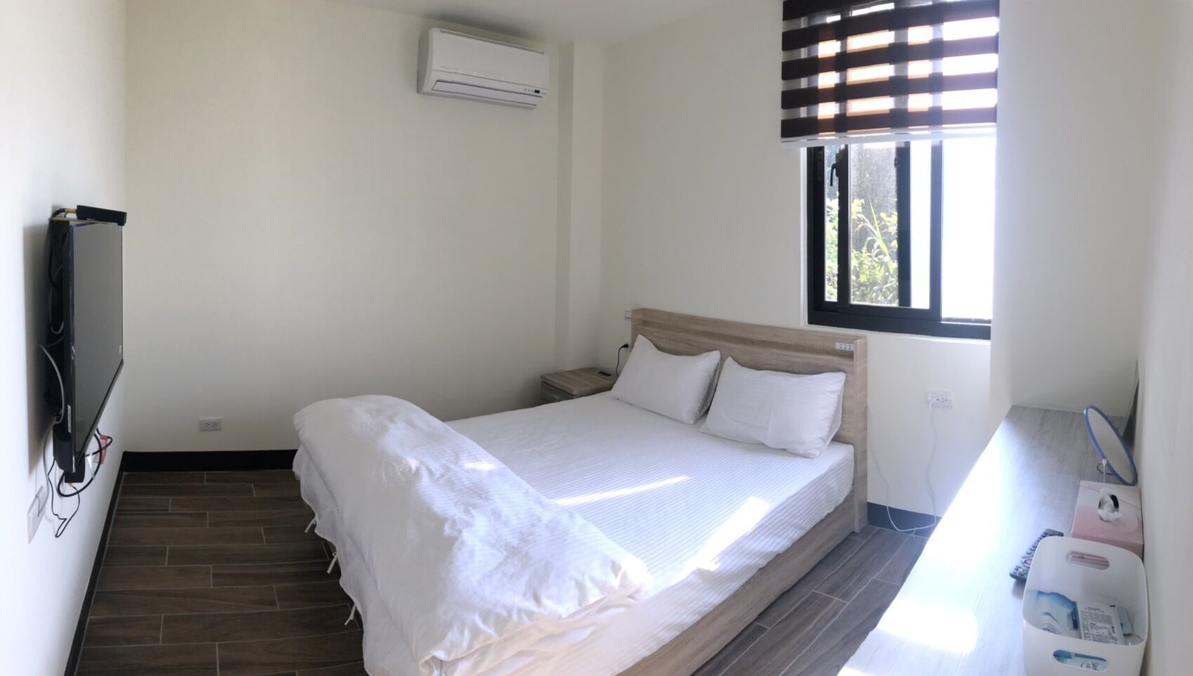 A small, clean room with a bed, a TV mounted on the wall, a window with blinds, and a small table with a mirror and a bottle.