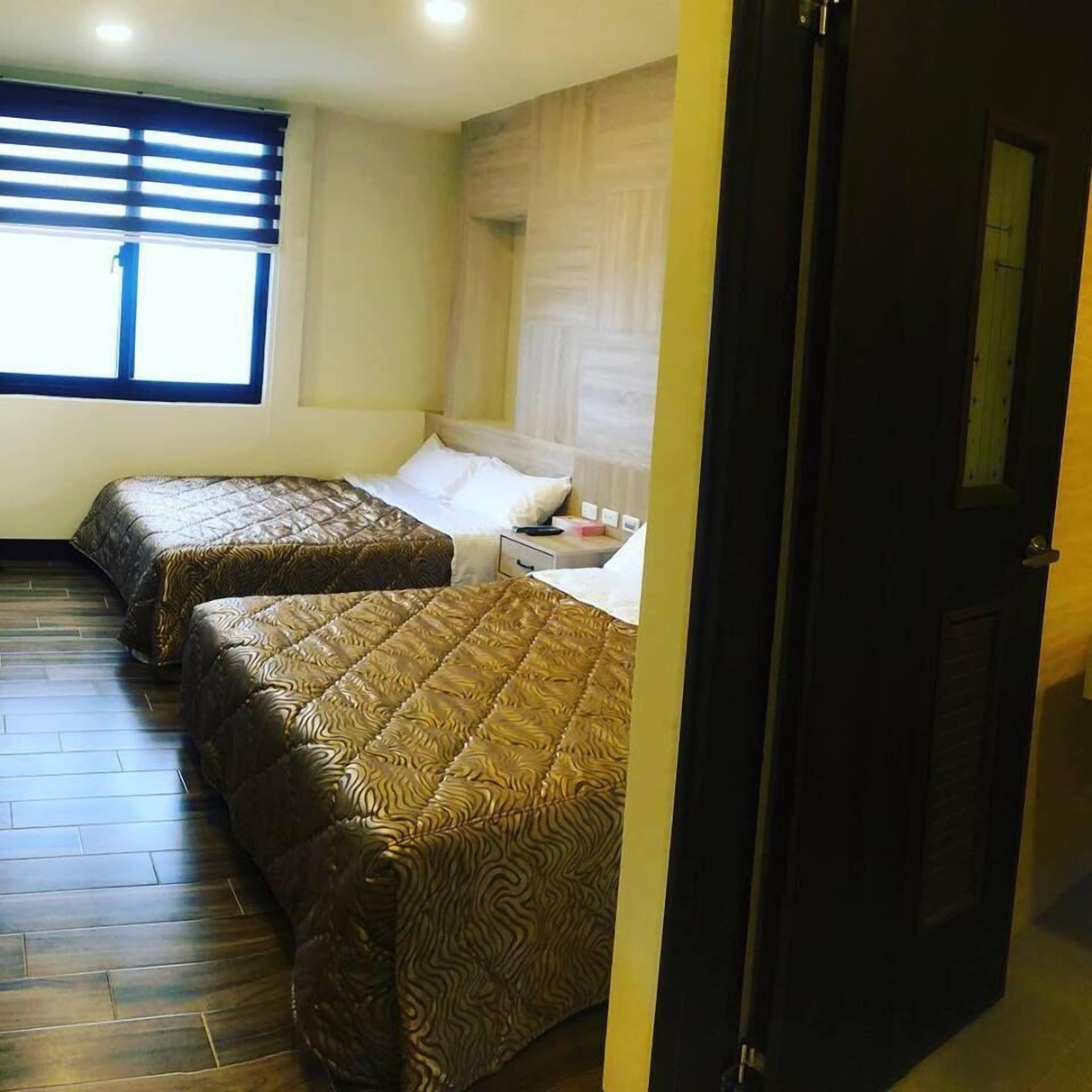 A room with two beds, wooden flooring, a window with blinds, and a door.