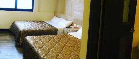 A room with two beds, wooden flooring, a window with blinds, and a door.