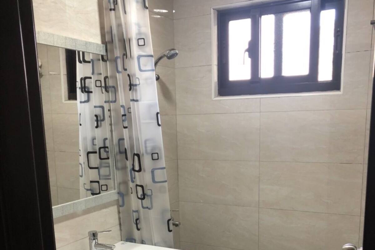 Un bany amb un lavabo blanc, una dutxa amb cortina estampada i una finestra amb persianes.