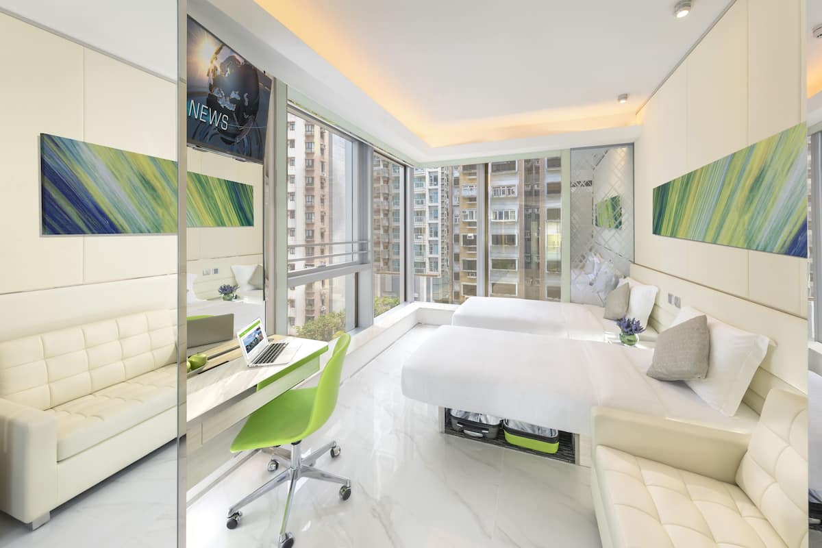 A modern hotel room with a large bed, a desk with a laptop, a green chair, and large windows offering a view of high-rise buildings.