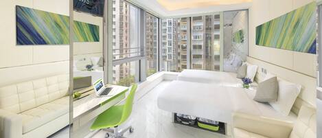 A modern hotel room with a large bed, a desk with a laptop, a green chair, and large windows offering a view of high-rise buildings.