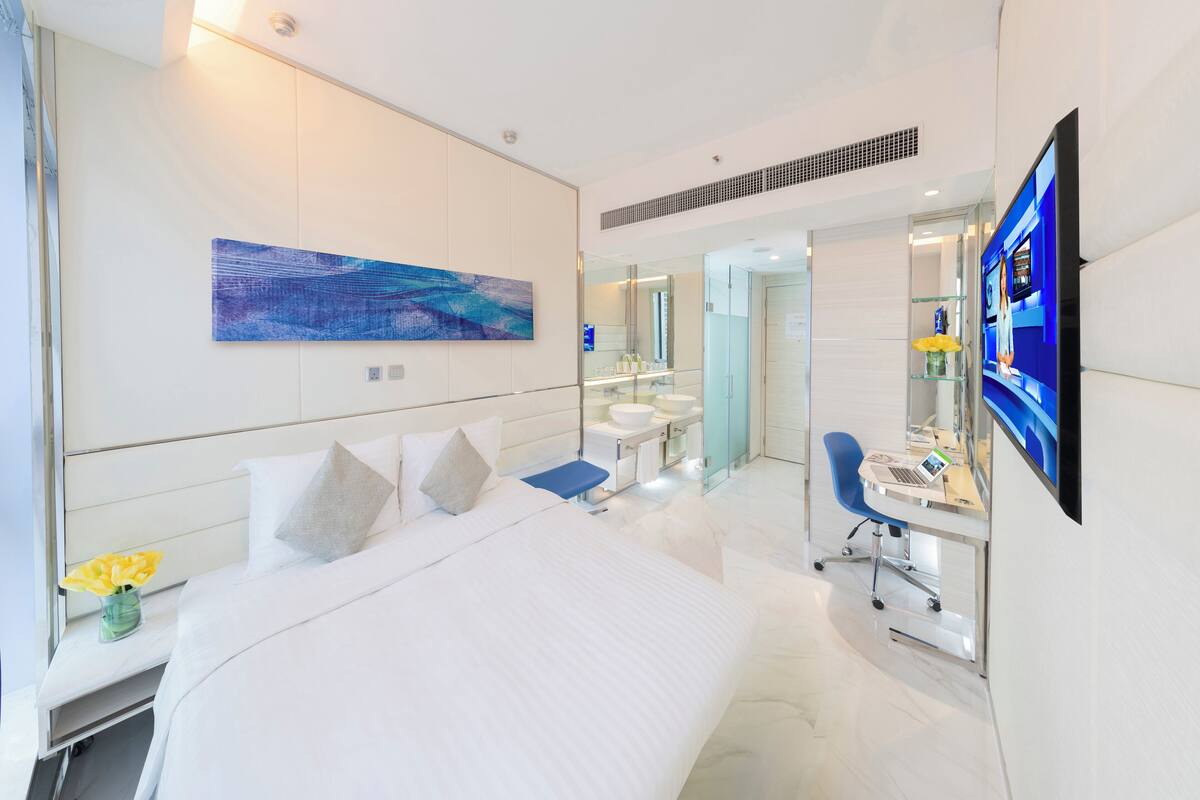A modern hotel room with a large bed, a desk with a laptop, a television, and a bathroom with a mirror and sink.