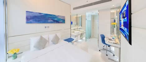 A modern hotel room with a large bed, a desk with a laptop, a television, and a bathroom with a mirror and sink.
