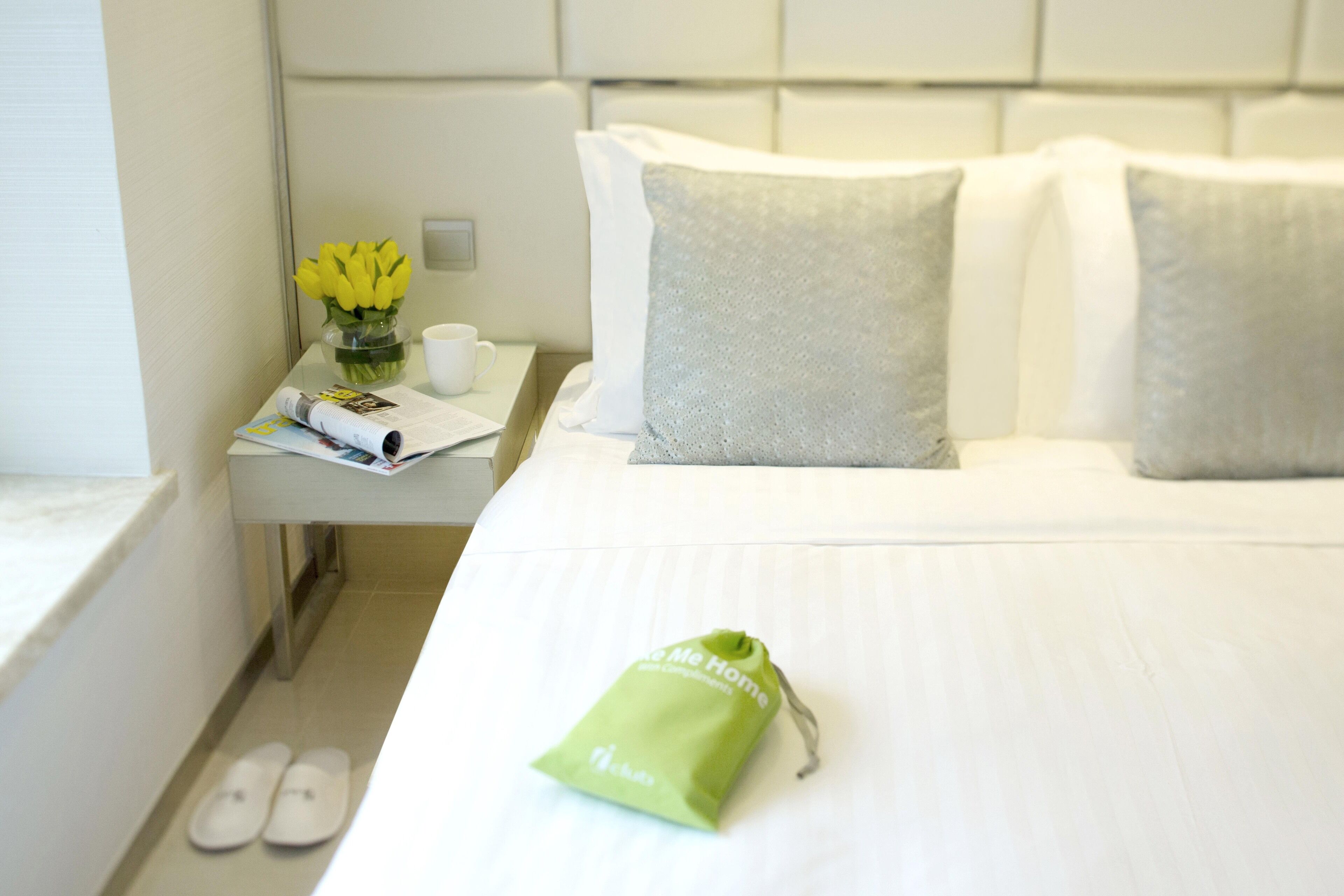 A hotel room with a neatly made bed, a nightstand with a vase of yellow flowers, a cup, and a magazine.