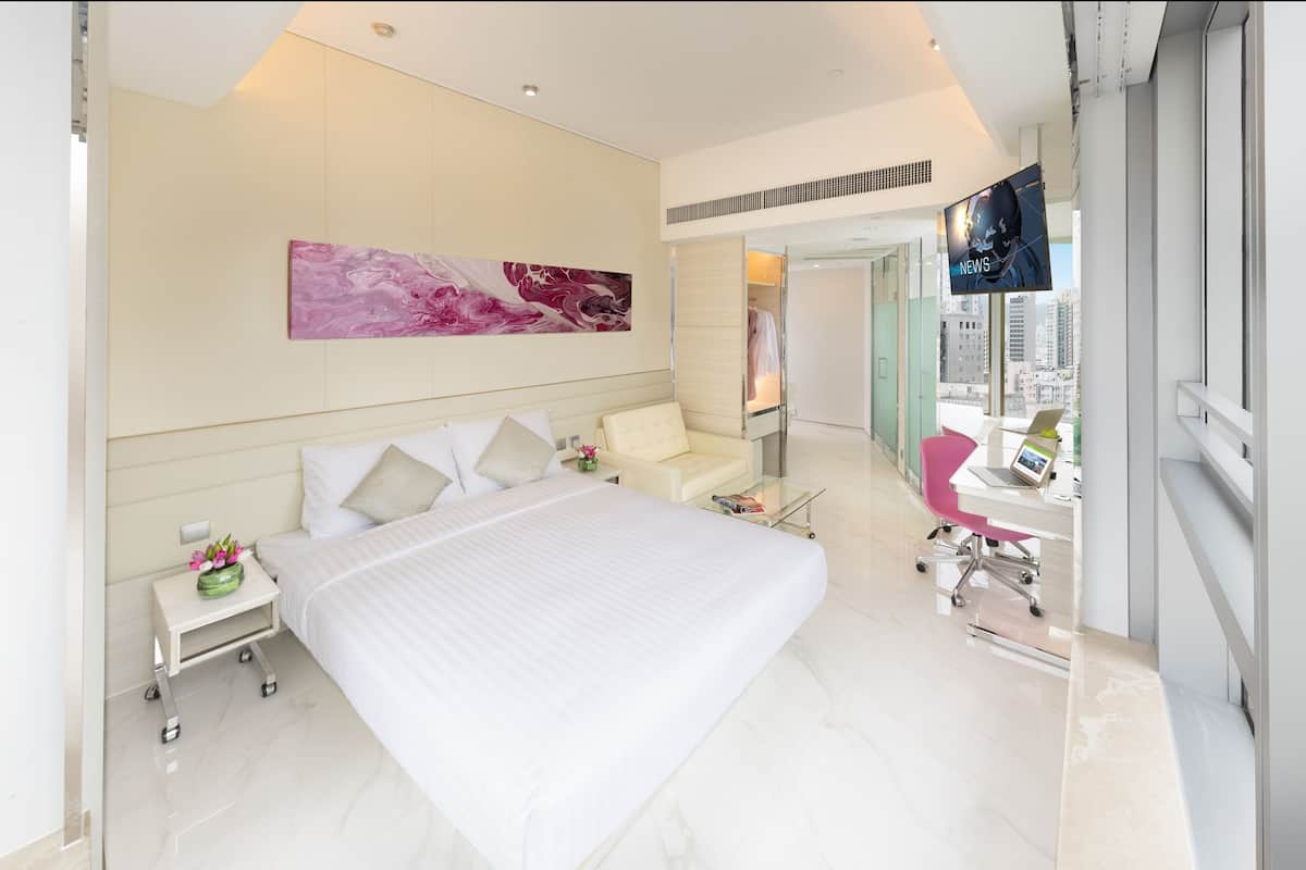 A modern hotel room with a large bed, a desk with a laptop, a pink chair, and a city view.