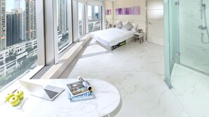 Un dormitorio moderno con una cama grande, ducha con cerramiento de vidrio y un portátil sobre una mesa de mármol.