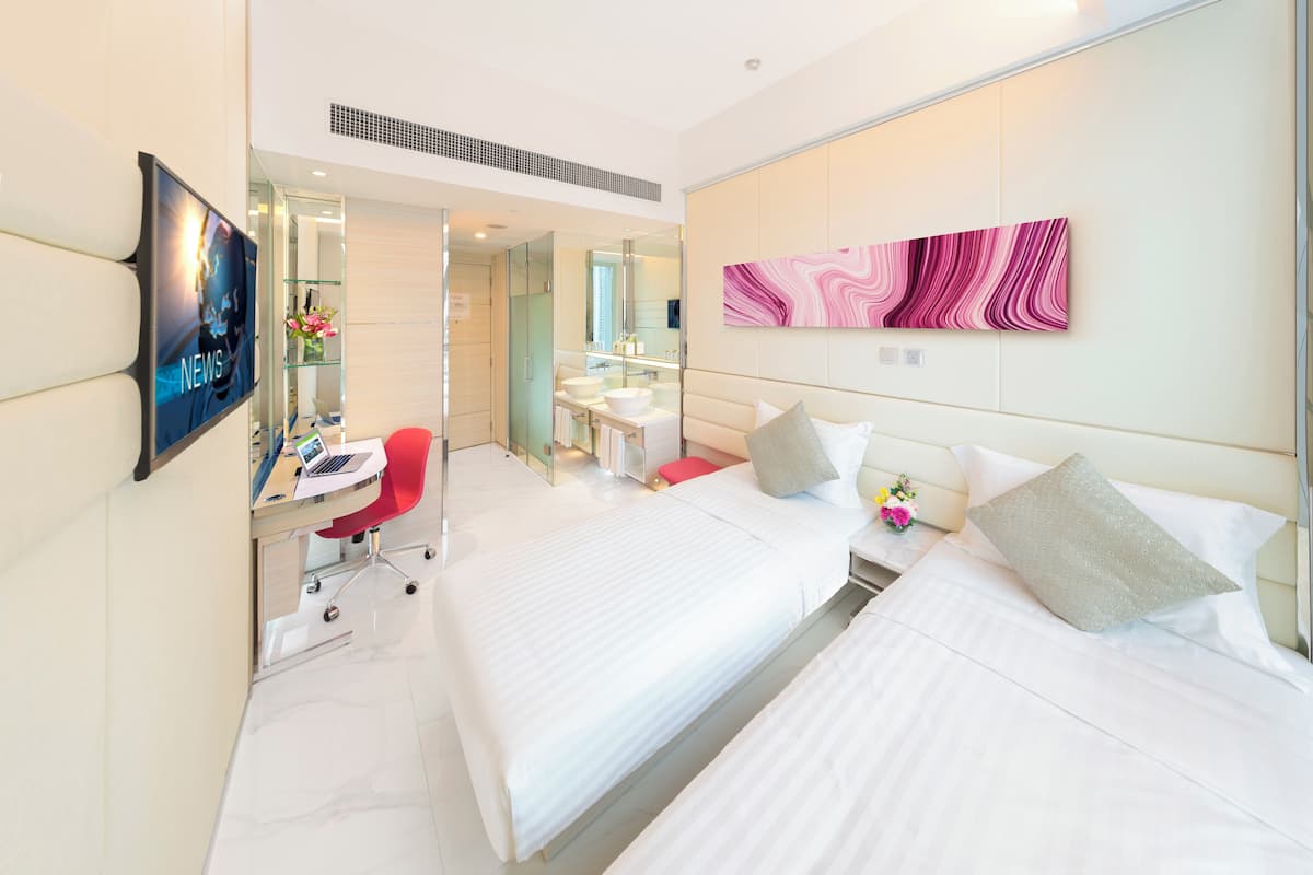 A hotel room with two beds, a desk with a laptop, a TV, a bathroom with a mirror, and a pink abstract painting on the wall.