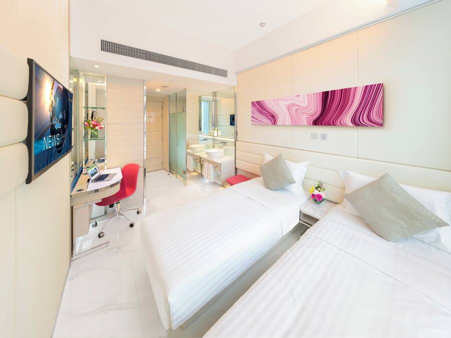 A hotel room with two beds, a desk with a laptop, a TV, a bathroom with a mirror, and a pink abstract painting on the wall.