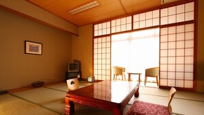 Ein traditioneller japanischer Raum mit Tatami-Boden, einem niedrigen Holztisch, zwei Stühlen, einem Fernseher und einer Schiebetür mit Gittermuster.