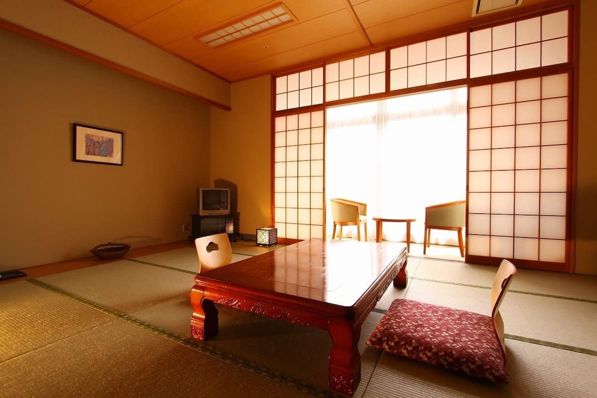 Una habitación japonesa tradicional con suelo de tatami, una mesa de madera baja, dos sillas, un televisor y una puerta corrediza con un diseño de cuadrícula.