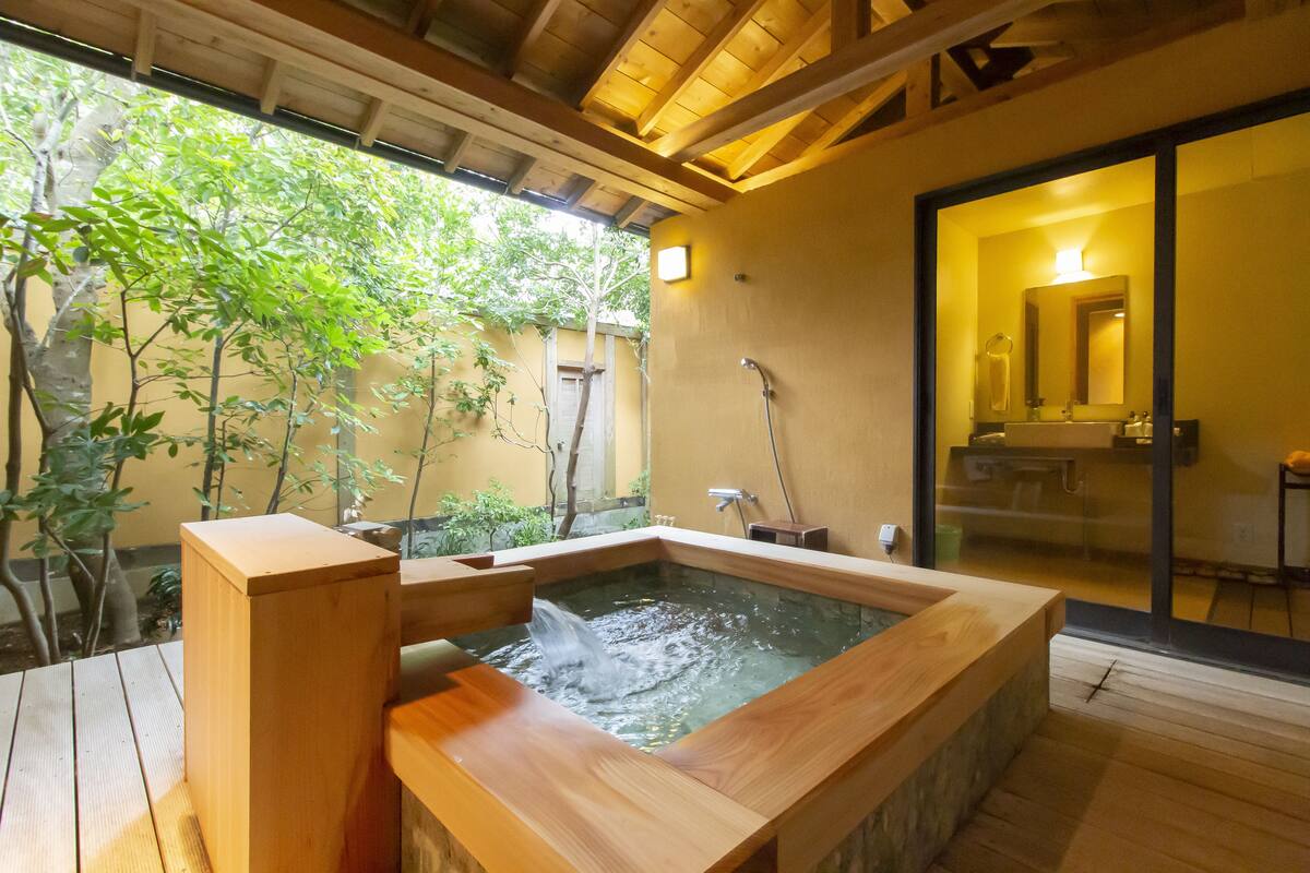 Tinaja de madera con agua corriendo, rodeada por una terraza de madera y un vidrio que da a un baño con espejo y lavamanos.
