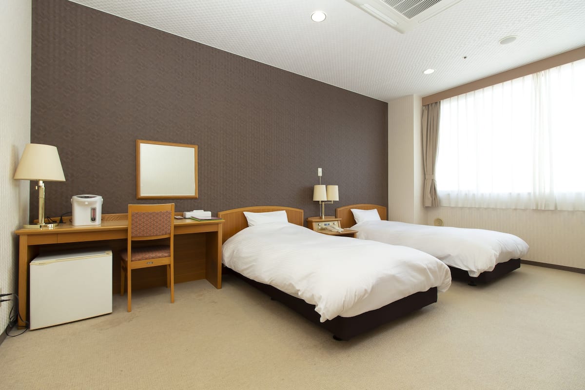 Habitación de hotel con dos camas, un escritorio, una silla, un refrigerador pequeño y una ventana con cortinas.