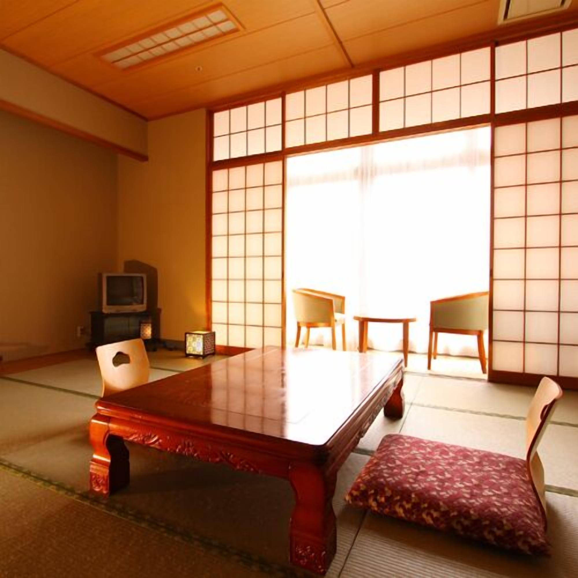 Um quarto japonês tradicional com tatames, uma mesa de madeira baixa, duas cadeiras e uma televisão.